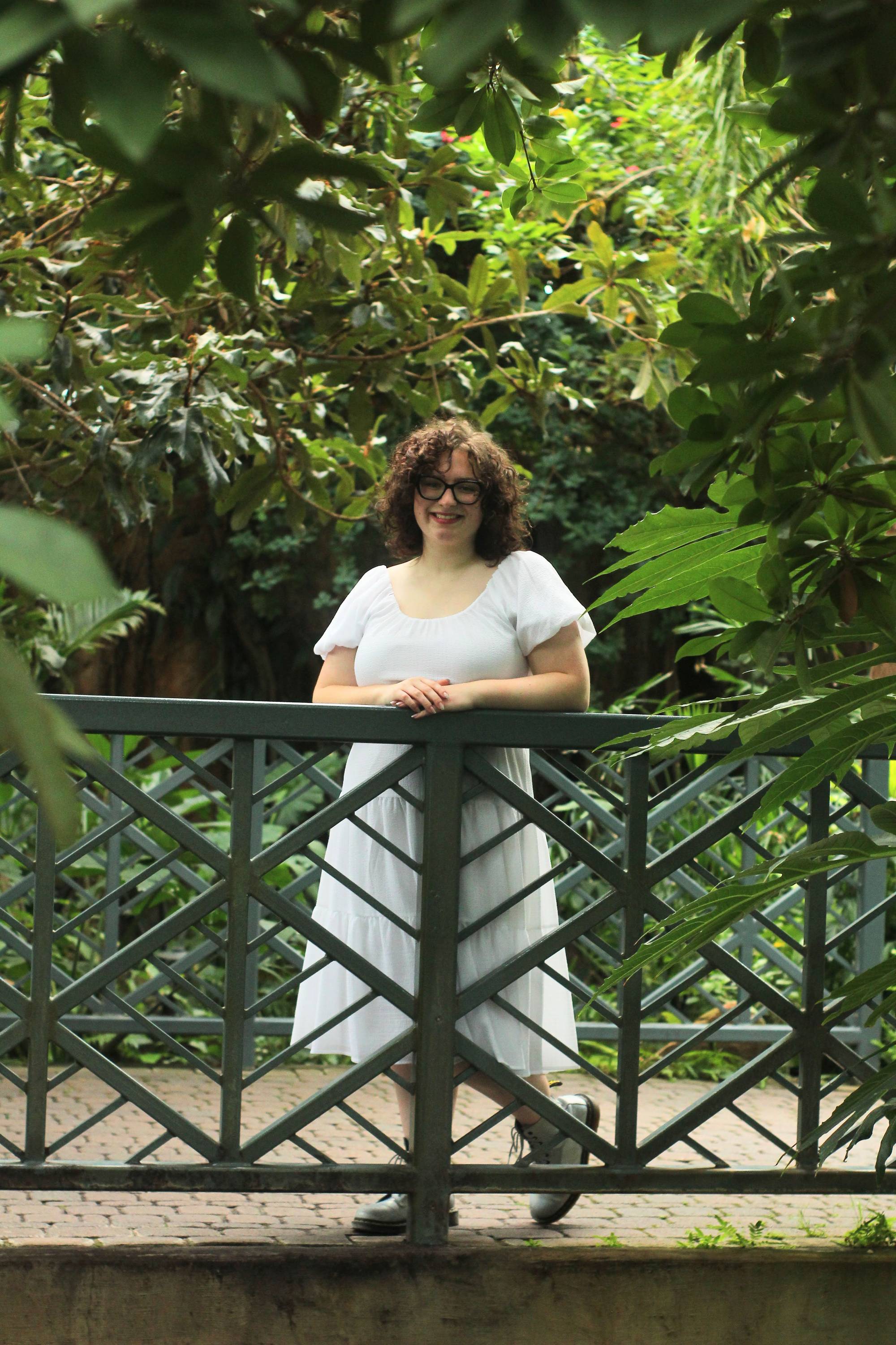 Woman with curly brown hair standing on a bridge surrounded by green foliage